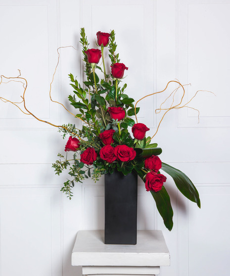 The tower of 12 roses dances with twigs of curly willow and textural greenery in a contemporary matte black ceramic vase. 