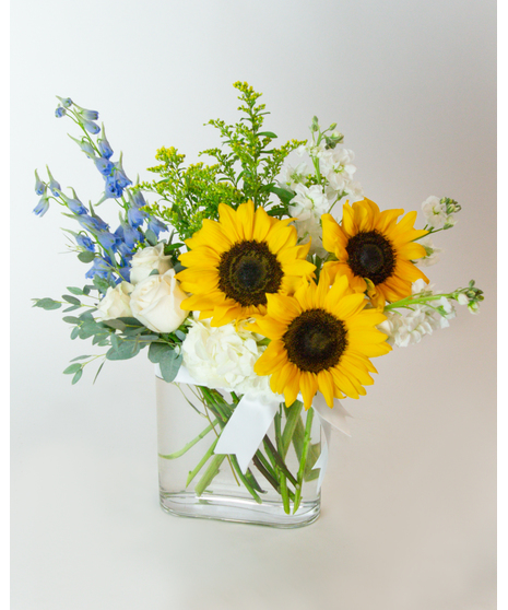 A bouquet of sunflowers, white roses, blue delphiniums, and greenery sits in a clear square glass vase, arranged against a plain white background.