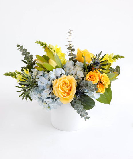 This arrangement has a lemon-lime color palette, with pastel blue hydrangea, baby blue eucalyptus, and vibrant yellow florals carefully arranged in a white cylinder.