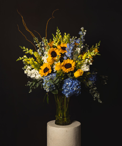 A large arrangement of sunflowers, hydrangea, delphinium, and snapdragons in a tall elegant glass vase. 