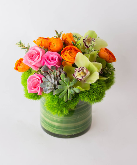 A floral arrangement in a glass vase features pink roses, orange ranunculus, green orchids, succulents, and green foliage, set against a plain white background.