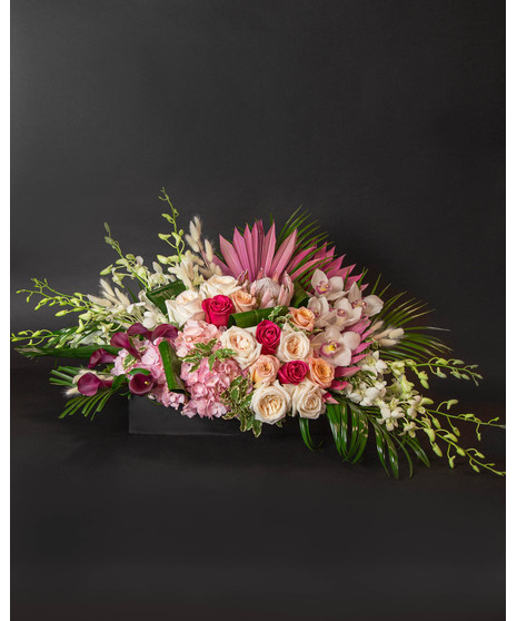A large and lush long and low arrangement of roses, palms, cymbidium and dendrobium orchids, hydrangea and calla lilies in an elegant black ceramic  vase. 