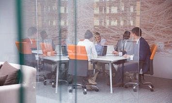A group of five professionally dressed individuals sit at a conference table, engaging in discussion with laptops and documents, in a modern office with glass walls and abstract wall art.