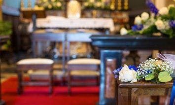A small bouquet of flowers sits on a pew in the foreground of a church, with an altar and two chairs in the background, and a red carpet in the aisle.