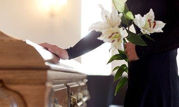 A person in black attire holds white lilies, resting one hand on a closed wooden casket, within an illuminated room with soft lighting and light-colored walls.
