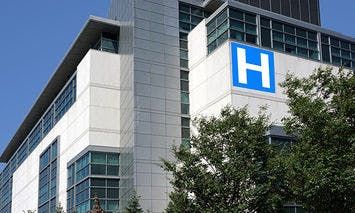 A modern hospital building stands tall with large windows. A prominent blue sign with a white 