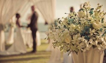 Flower arrangement in foreground; two people stand, likely in conversation, under a white canopy in the lawn. Blurry background with focus on the floral bouquet.